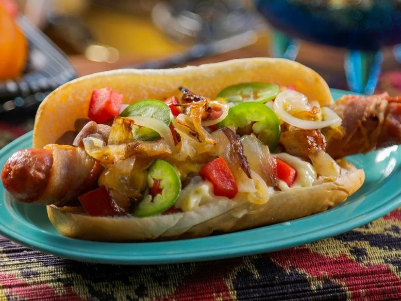 Loaded hot dog with grilled onions, peppers, and tomatoes on a colorful plate.