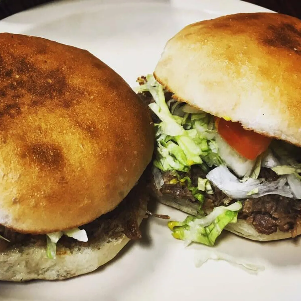 Two homemade burgers with lettuce and tomato on white plate.